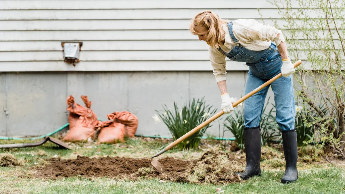 Woman raking soil where lawn meets bare dirt beside a house
